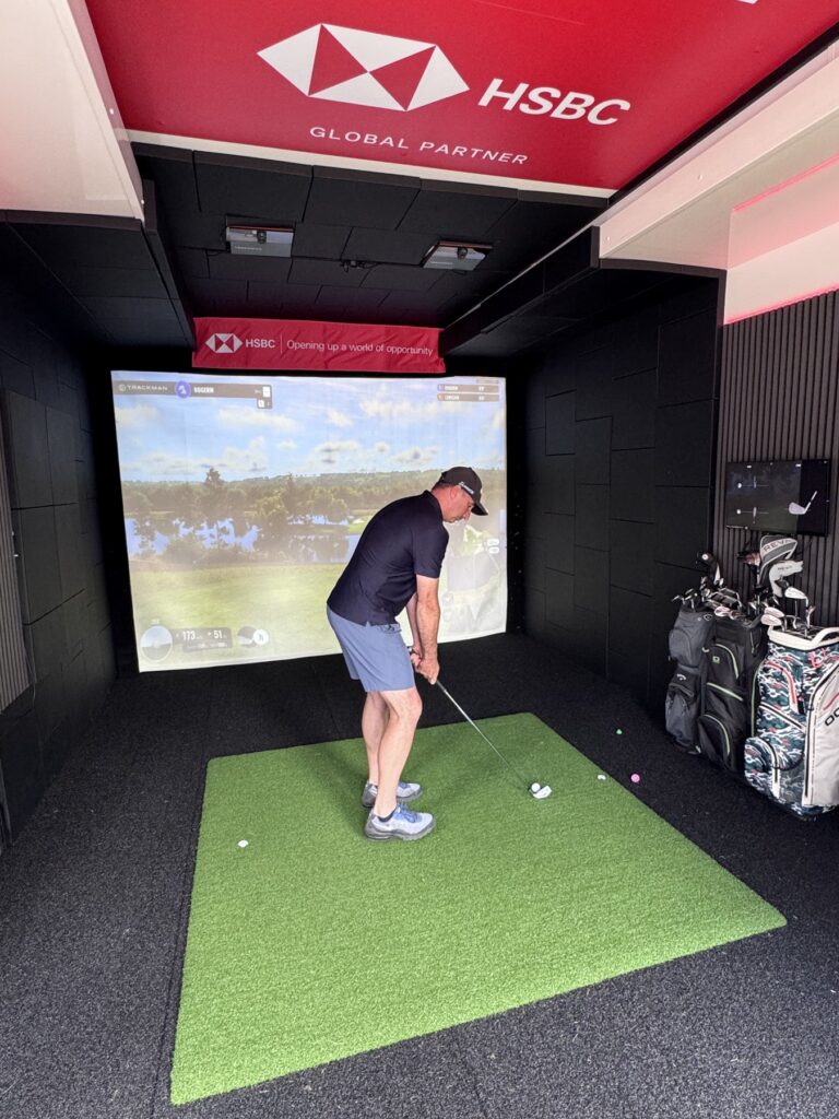 Golfer mid-swing inside the HSBC branded Dryvebox golf simulator on a TrackMan hitting mat