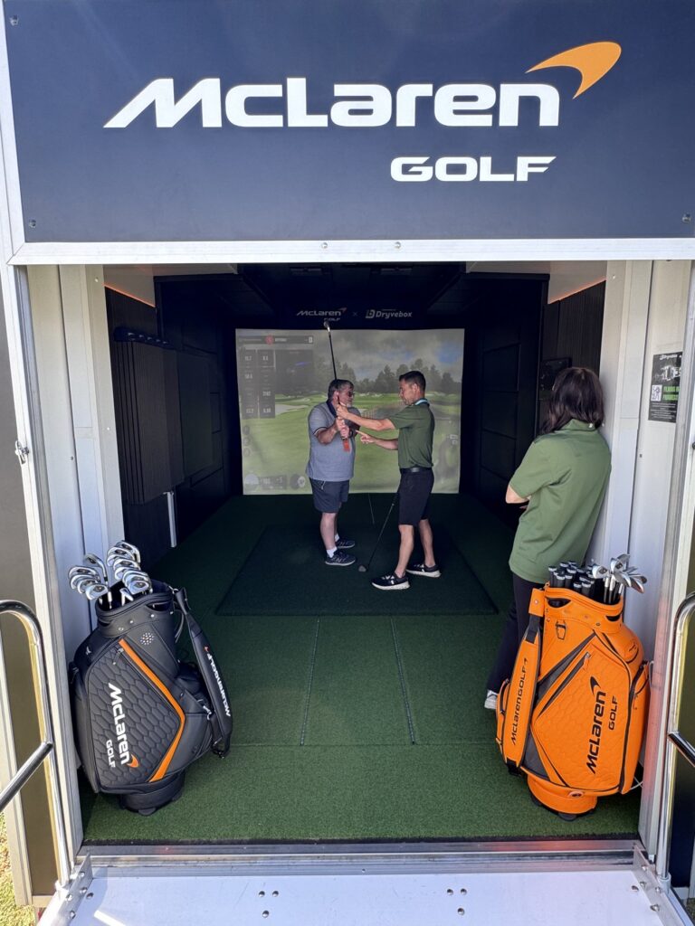 Two golfers playing inside the McLaren Golf branded Dryvebox mobile golf simulator at a UK event